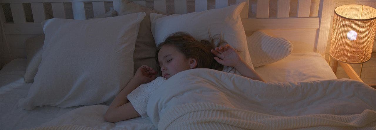 Child sleeping in bed with white sheets and bedside lamp