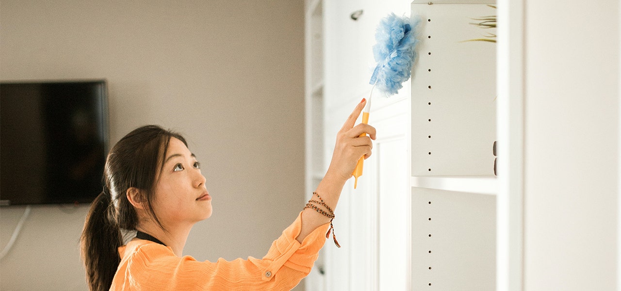 Woman using a duster to clean shelves in her home