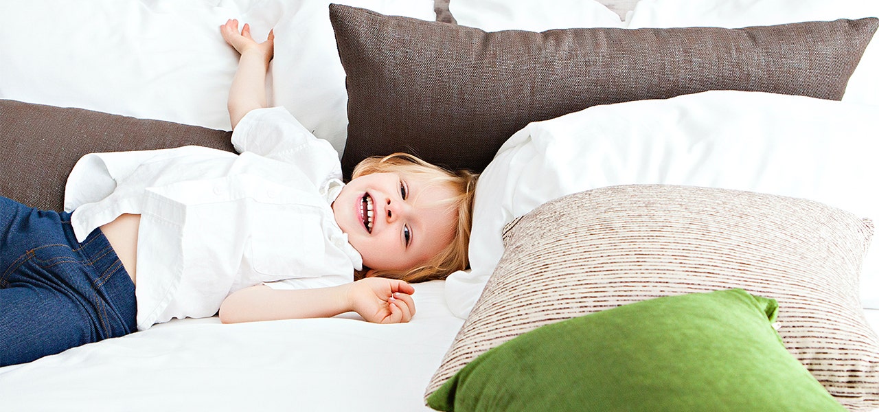 Toddle lying back on a big kid bed with lost of pillows and smiling